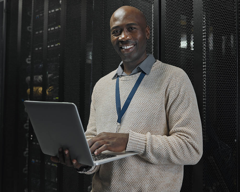 A person standing in a server room holding a laptop and smiling.