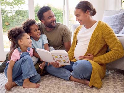 Four people, two children and two adults, smiling and sitting on a living room floor. A pregnant adult is holding a children's book.